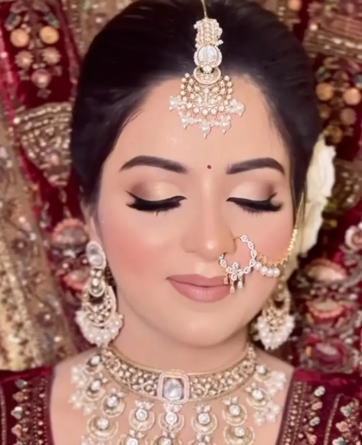 A close-up of a bride with soft gold eye makeup and a nude lip, wearing a pearl maang tikka, a pearl nath and a wide kundan choker, a maroon embroidered lehenga behind her.