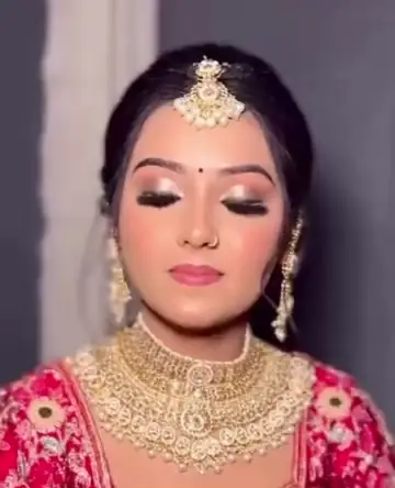 A bride in a red and pink lehenga with a layered gold choker, long gold earrings and a maang tikka, eyes lowered.