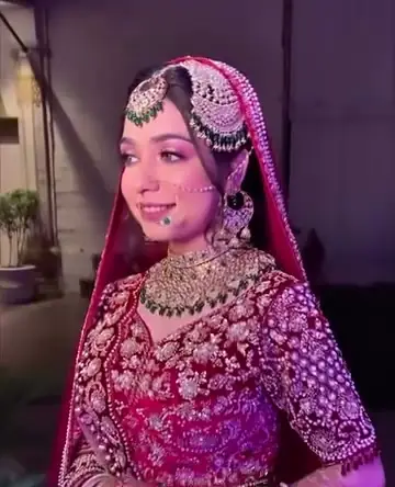 A bride in a magenta and pink lehenga with a pearl passa over one ear, a green and gold necklace and a nose ring joined by a chain.