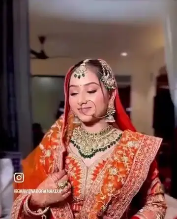 A bride in an orange and red lehenga with a green and gold necklace and maang tikka, looking down, indoors.