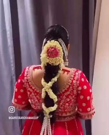 The back of a bride's long plait, dressed with a pink and white flower gajra, over a red embroidered blouse.
